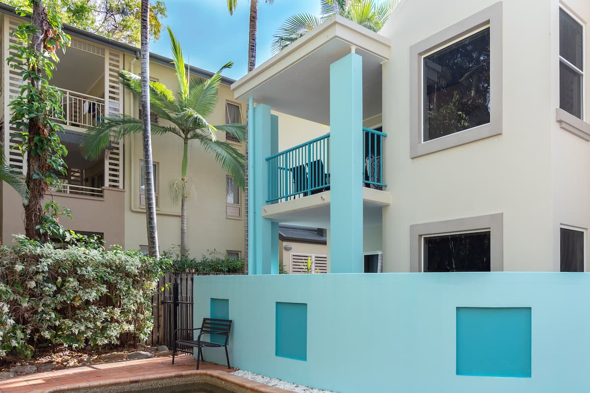 A modern building with a pool, featuring a light blue exterior, a balcony with blue railings, and a small garden area with a bench.