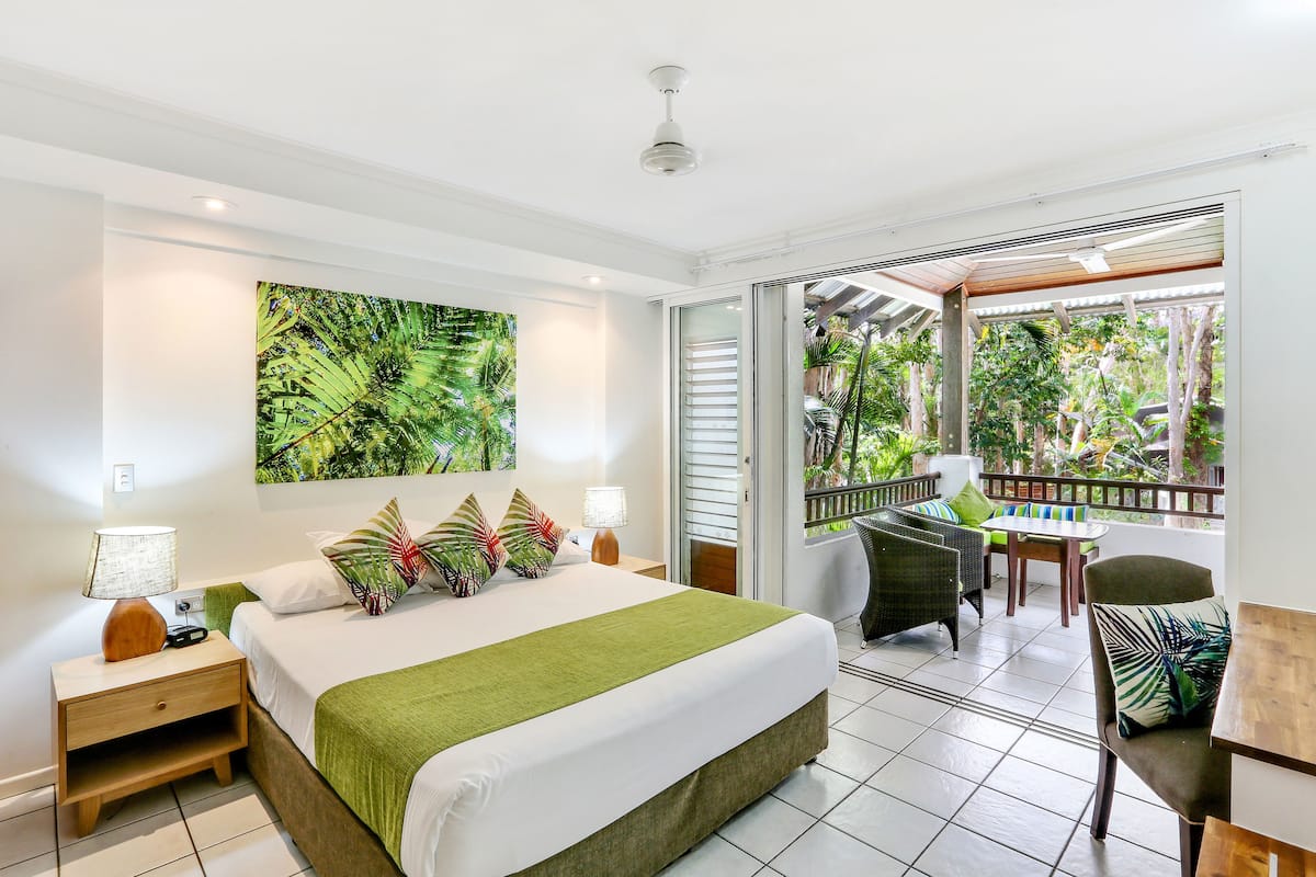 A hotel room with a bed, nightstands, a table, chairs, and a balcony with a view of greenery.