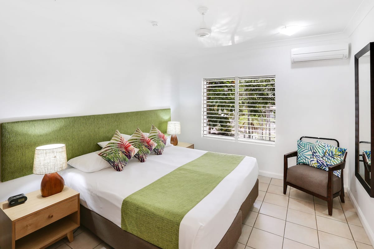 A neatly arranged hotel room with a bed, bedside table, lamp, chair, window with blinds, and an air conditioning unit.