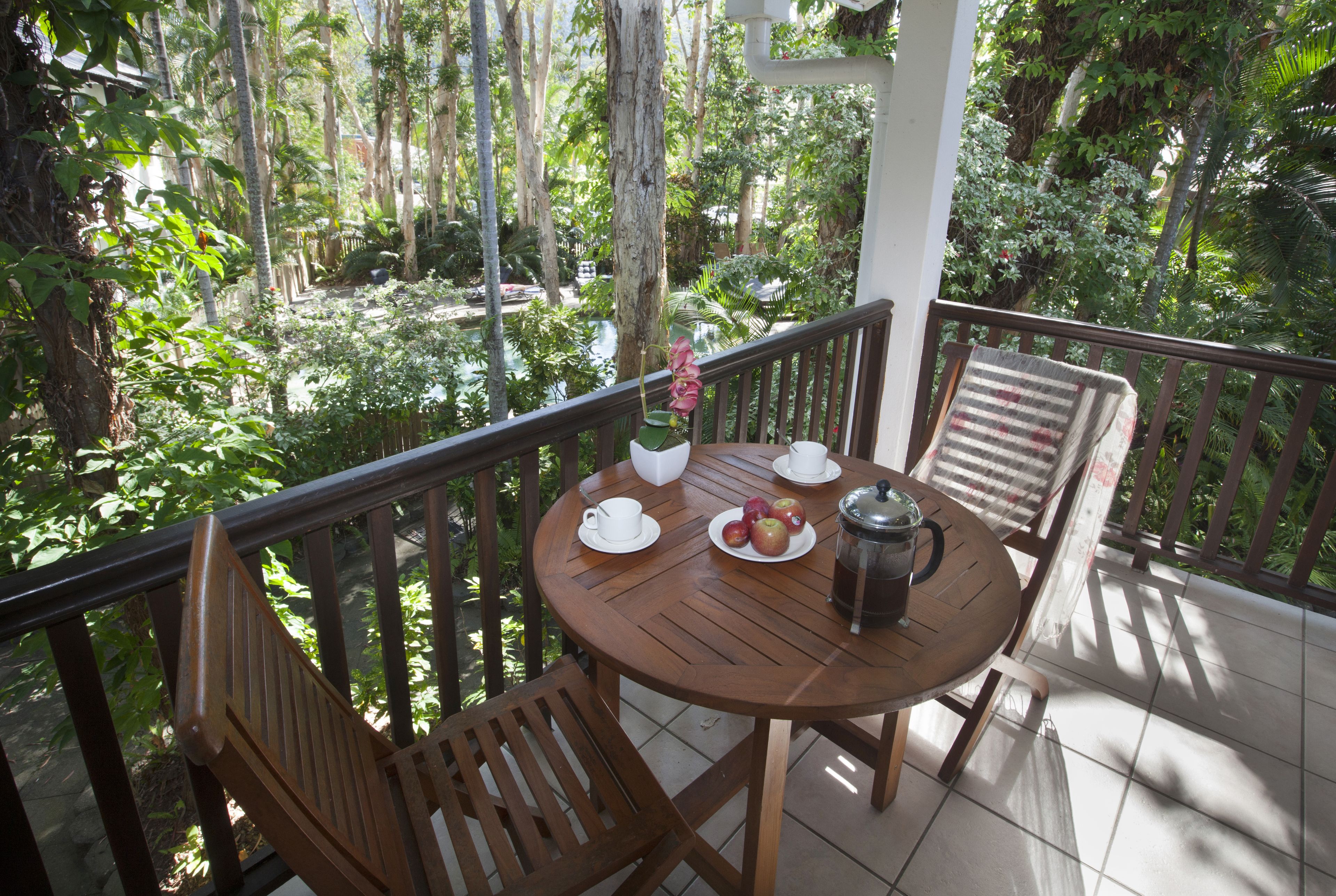 A wooden table with a coffee pot, cups, and apples on a balcony with a view of a lush garden.