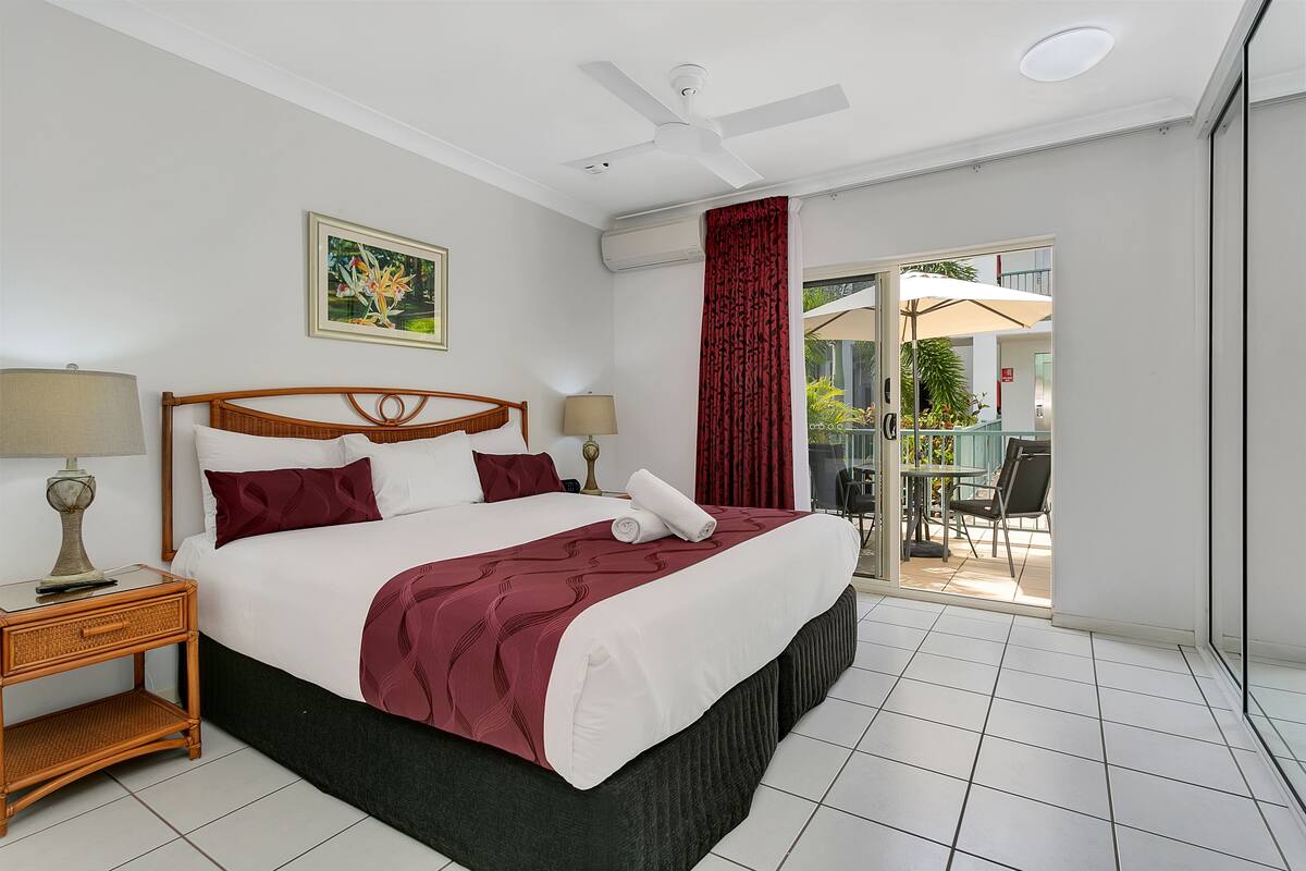 A hotel room with a large bed, two bedside tables with lamps, a ceiling fan, a window with red curtains, and a view of an outdoor patio.