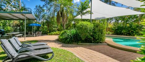 A poolside area with lounge chairs, a covered seating area, and a swimming pool surrounded by lush greenery.