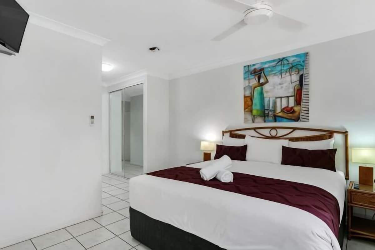 A hotel room with a bed, a painting on the wall, a ceiling fan, and a nightstand with a lamp.
