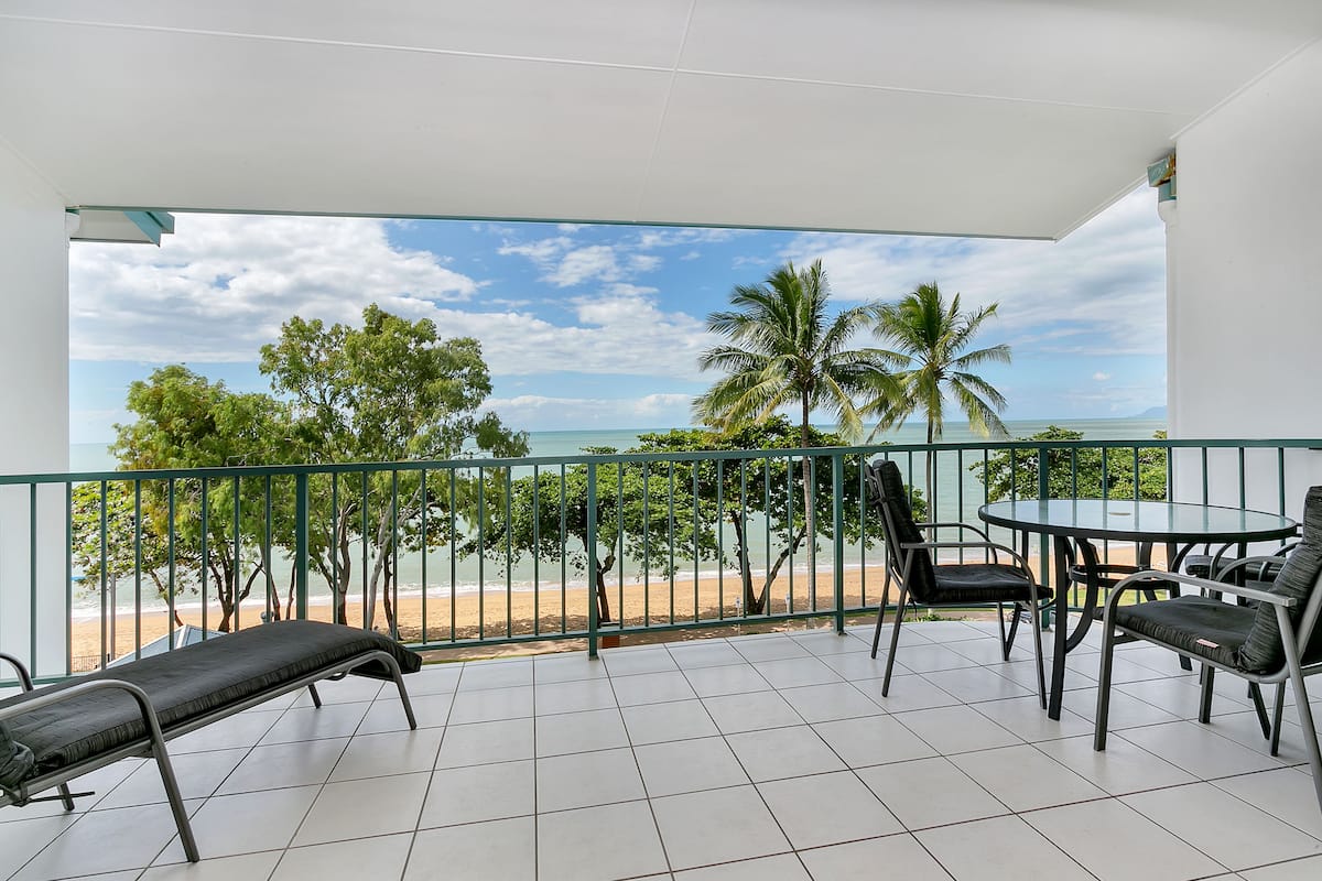 A balcony with a view of the beach, a table, and two chairs.