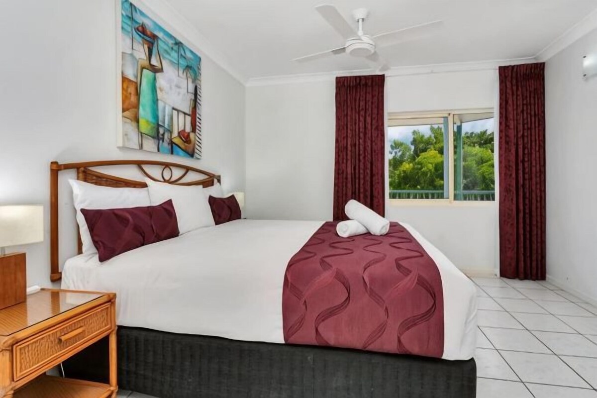 A hotel room with a bed, a nightstand, a ceiling fan, a window with curtains, and a painting on the wall.