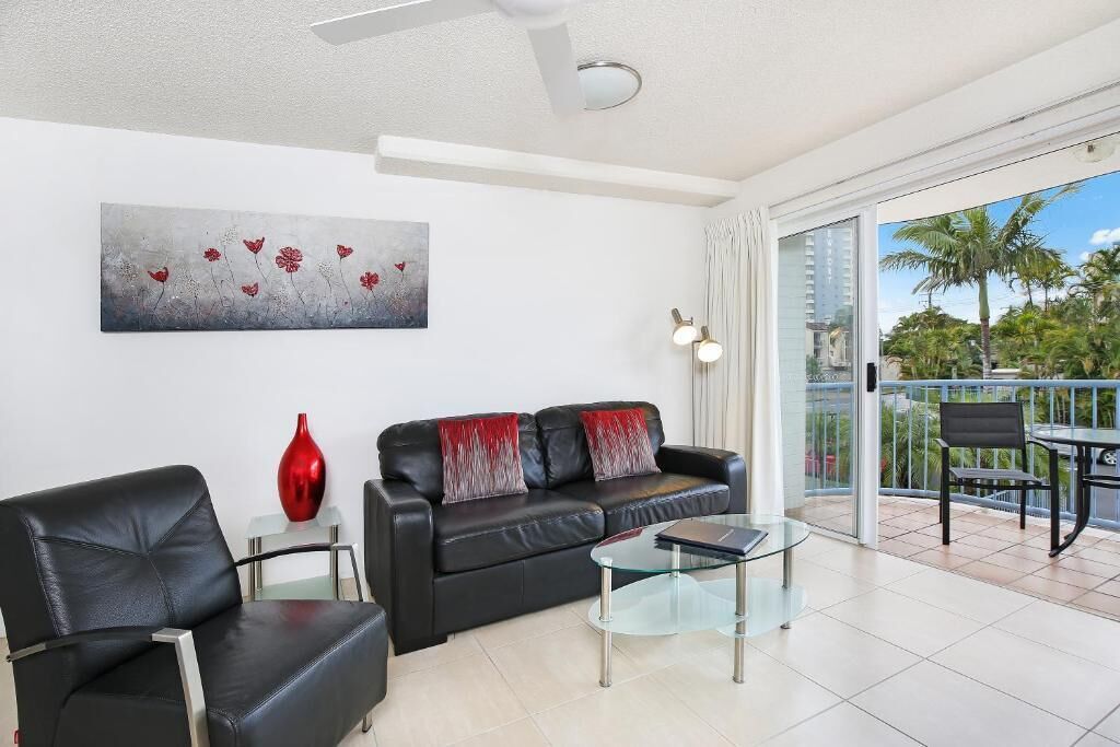 Un salon moderne avec un canapé en cuir noir, une table basse en verre et un vase rouge. Un tableau représentant des fleurs est accroché au mur, et un balcon offre une vue sur des palmiers.