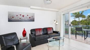 Un salon moderne avec un canapé en cuir noir, une table basse en verre et un vase rouge. Un tableau représentant des fleurs est accroché au mur, et un balcon offre une vue sur des palmiers.