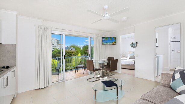 A modern living room with a glass coffee table, a sofa, and a dining area with chairs. A ceiling fan is visible, and there is a balcony with a view of palm trees and buildings. - Beachside Mooloolaba (Mooloolaba)