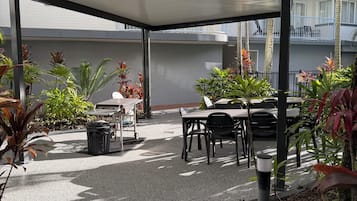 Un área de estar al aire libre con techo, mesas y sillas, rodeada de macetas con plantas y suelo de grava.