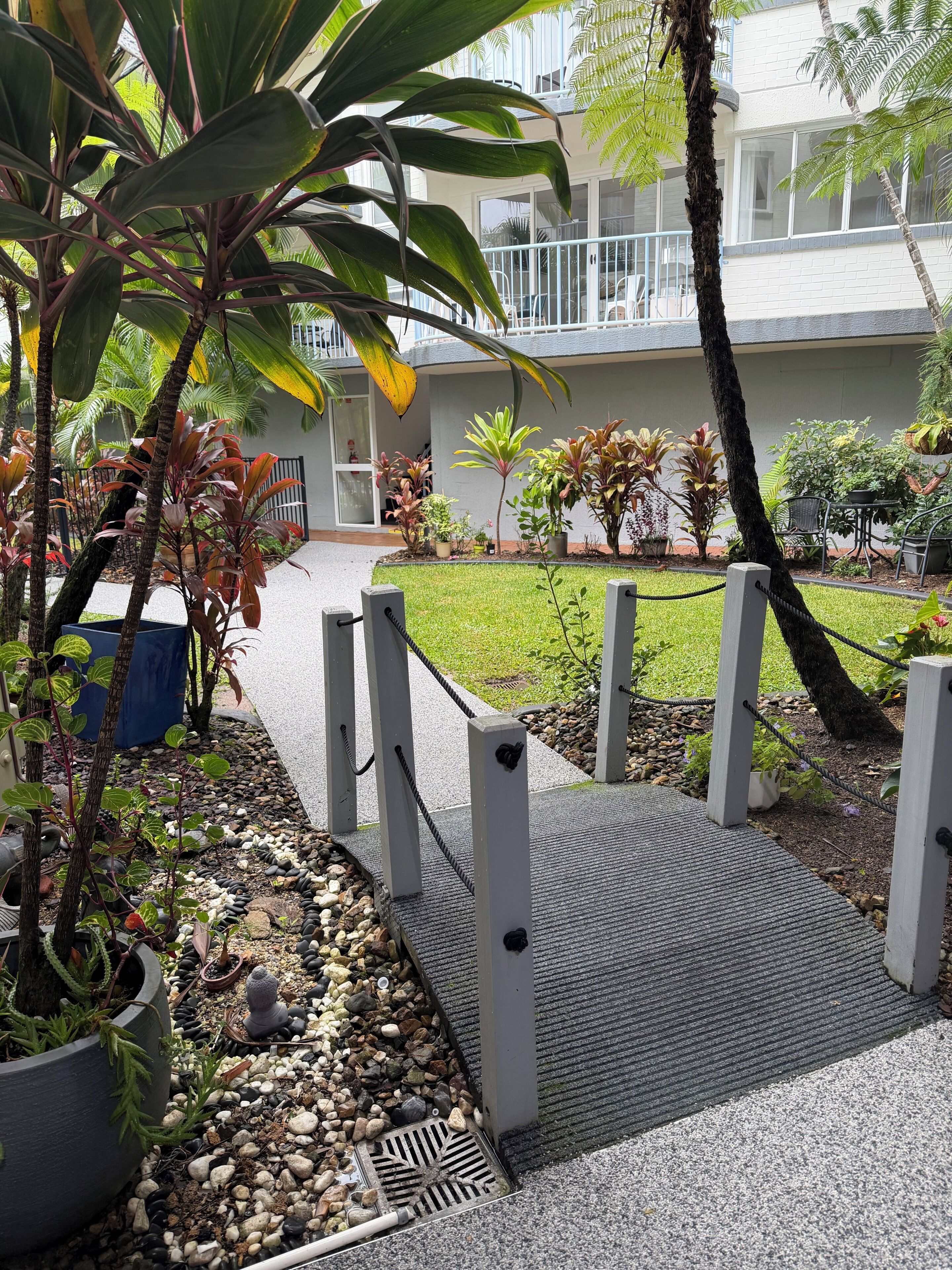 A pathway leading to a building with a balcony, surrounded by various potted plants and a small garden area.