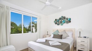 A neatly arranged bedroom with a large bed, a bedside table with a lamp, a wall art piece, and a view of palm trees through the window. - Beachside Mooloolaba (Mooloolaba)
