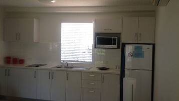 Una cocina con armarios blancos, microondas, refrigerador y ventana con persianas.