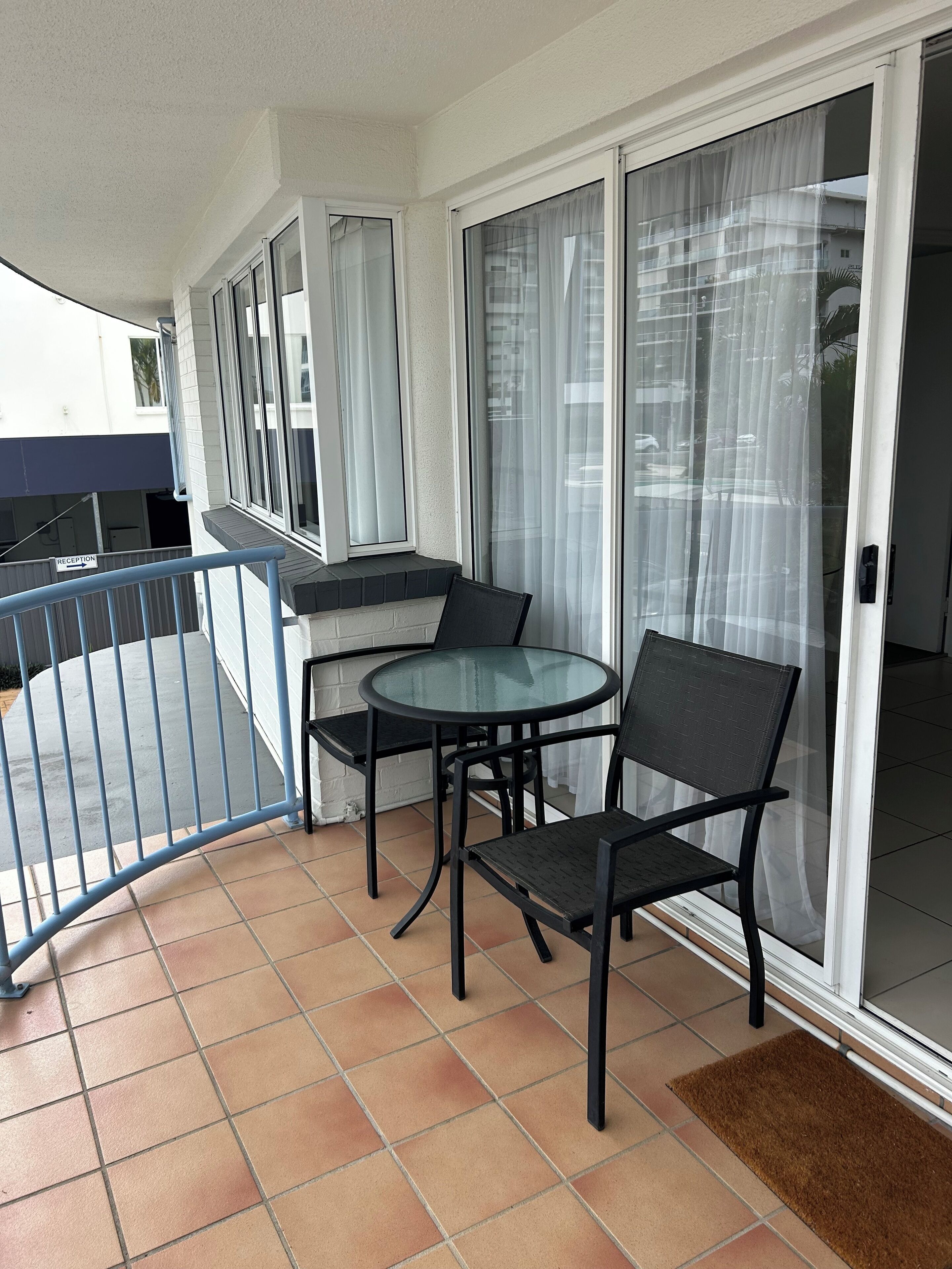 A balcony with a glass table and chairs, a blue railing, and a sliding glass door.