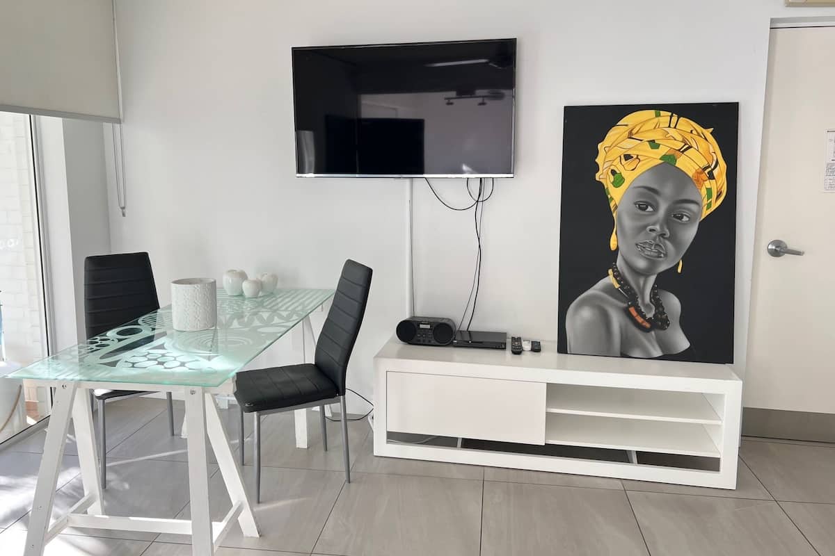 A modern living room with a glass dining table, black chairs, a white entertainment unit, a flat-screen TV, and a large abstract painting.