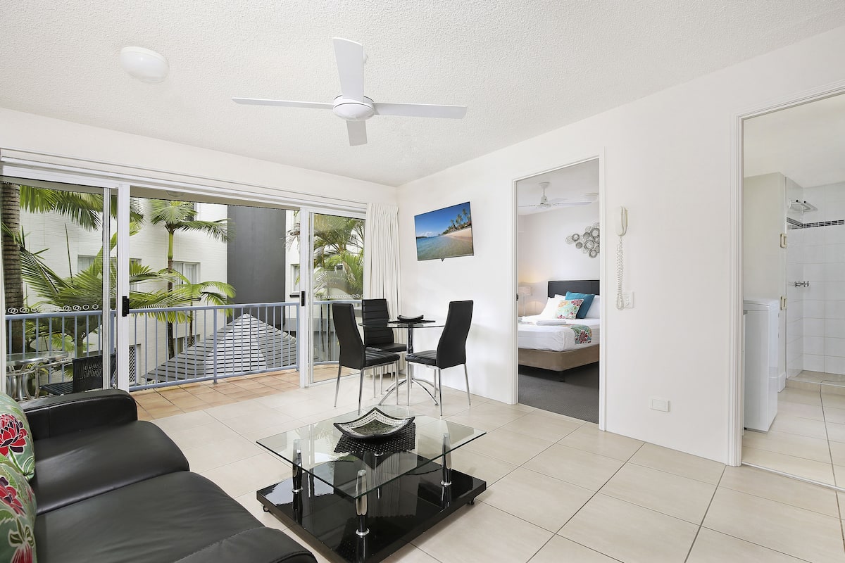 A modern living room with a glass coffee table, black sofa, and a ceiling fan. There is a balcony with a view of palm trees and a staircase leading to an upper level.
