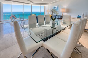 A modern dining area with a glass table and white chairs, overlooking a balcony with a view of the sea.