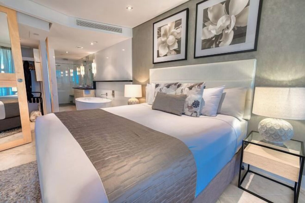 Una habitación de hotel moderna con una cama grande, una mesita de noche y un espejo. Hay cuadros enmarcados en la pared y una ventana con cortinas.