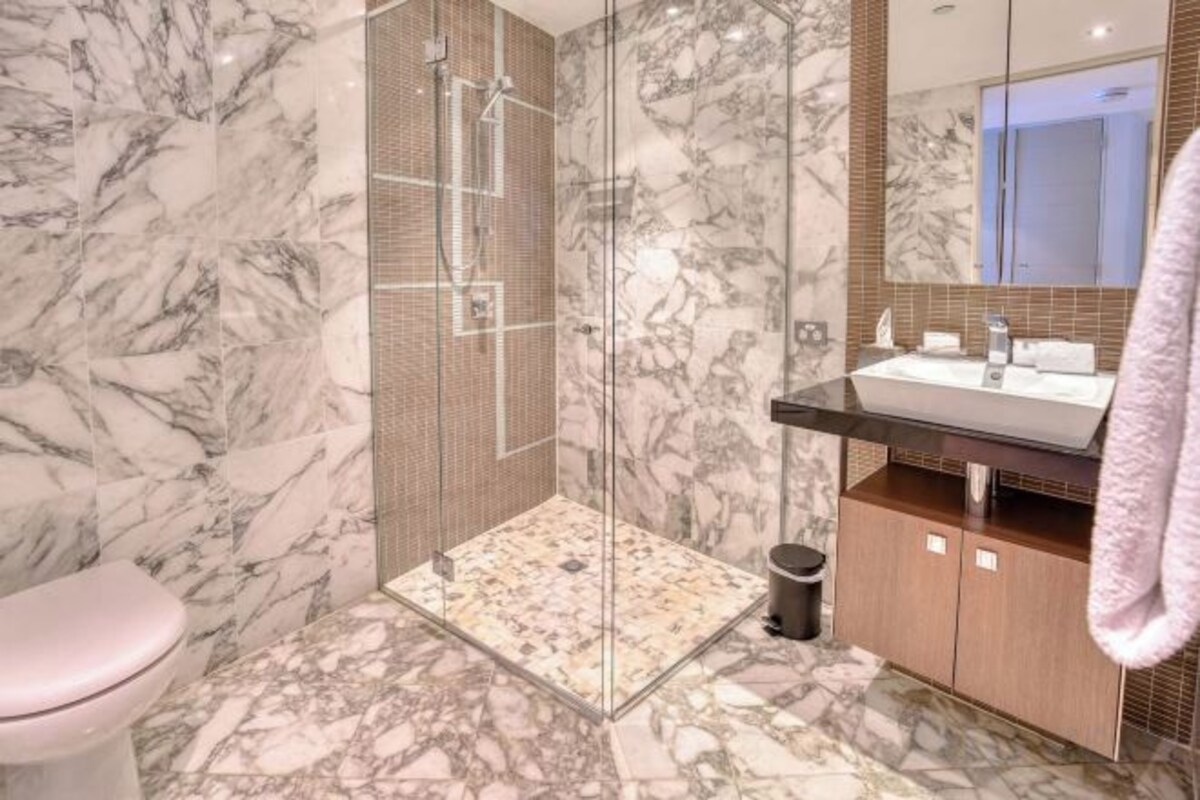 A bathroom with a glass-enclosed shower, a marble floor, a sink with a mirror, and a toilet.