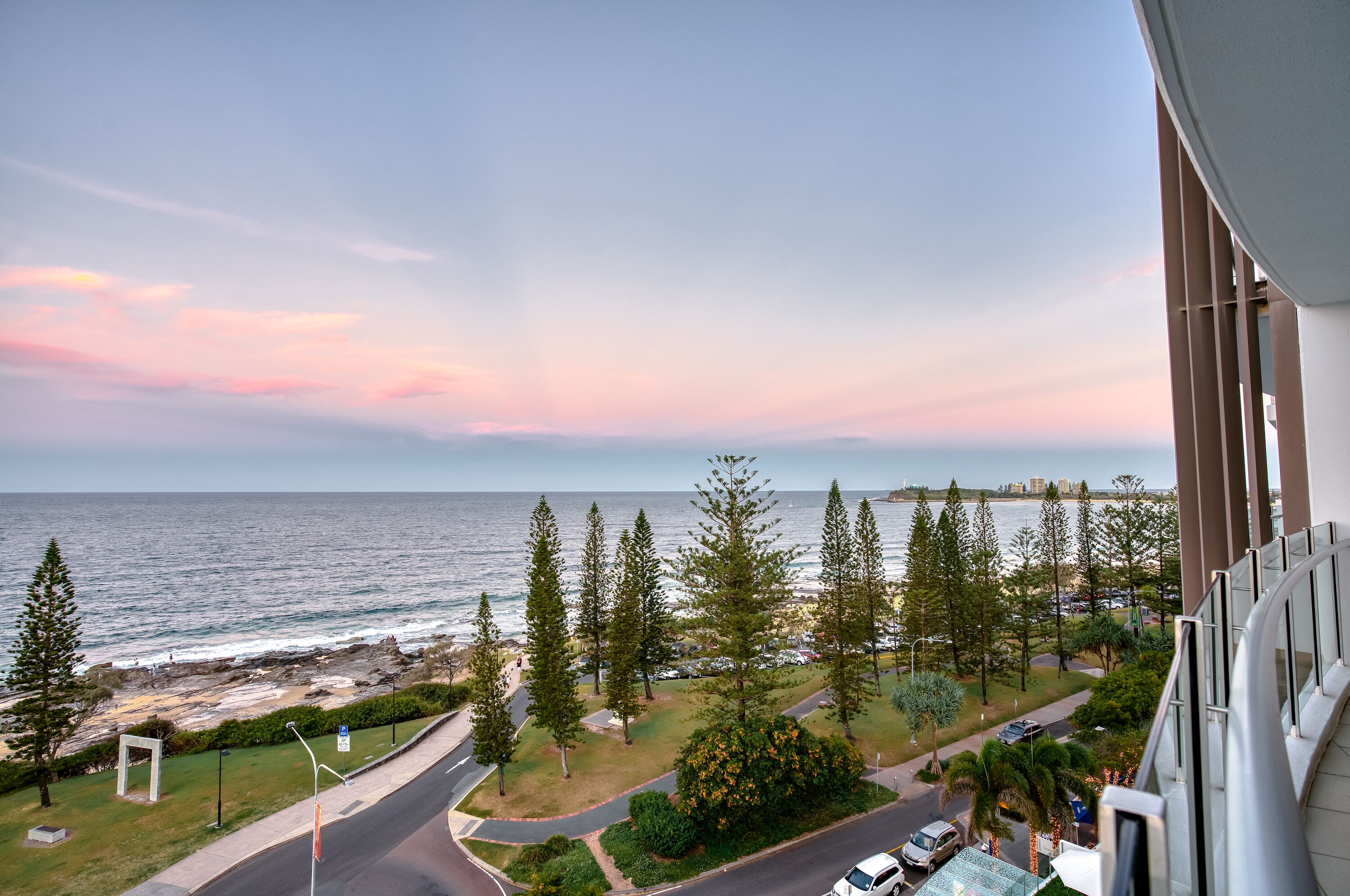 A coastal view from a balcony overlooking a beach with trees and a road.