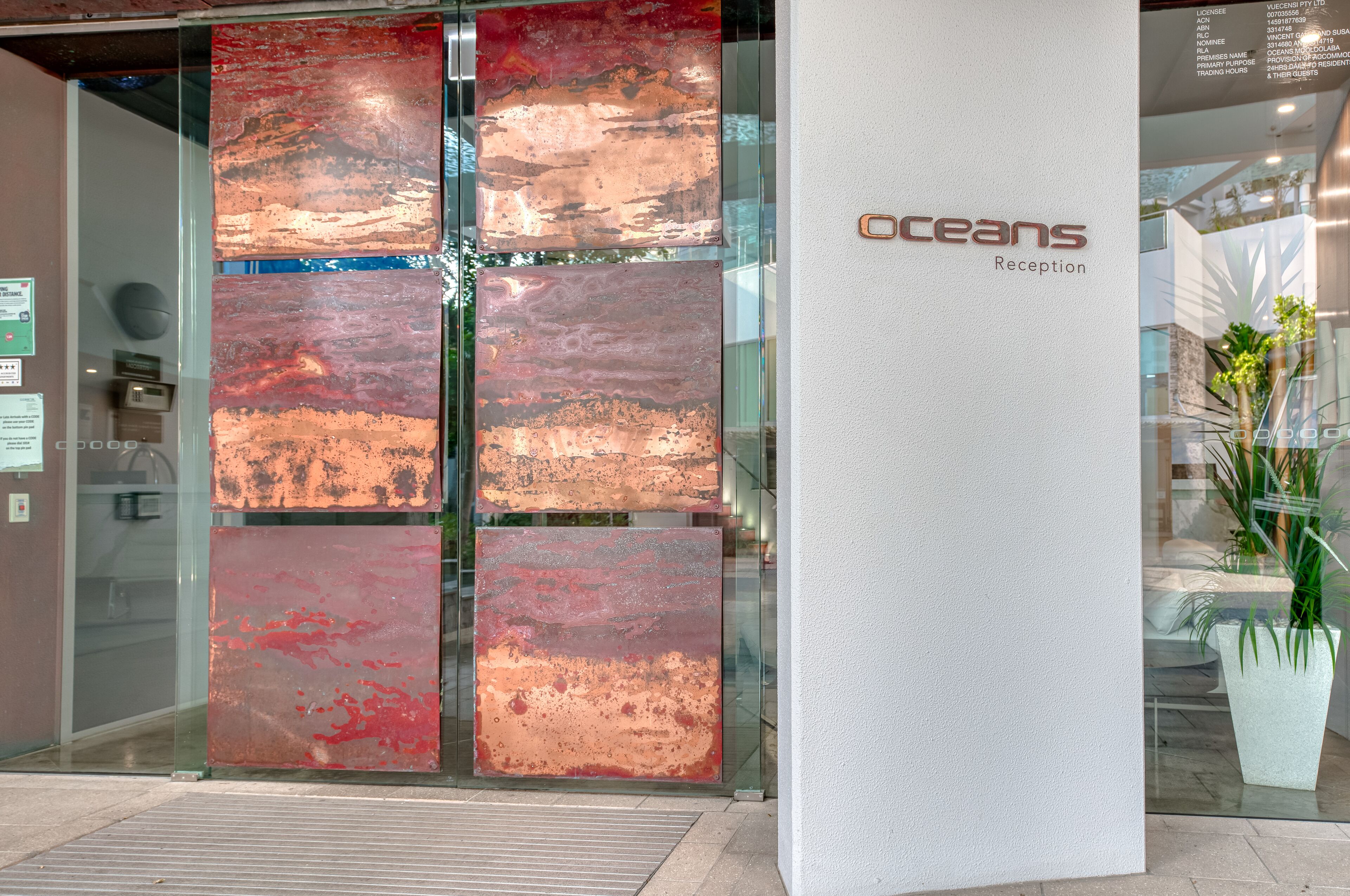 Zona de recepción con puerta de vidrio, un pilar con el nombre 'Oceans' y la palabra 'Recepción', y una planta en maceta a la derecha.