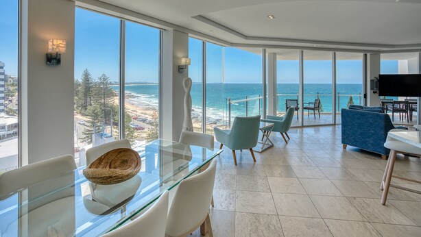 A modern living room with a glass table, white chairs, a blue sofa, and a large view of the ocean.