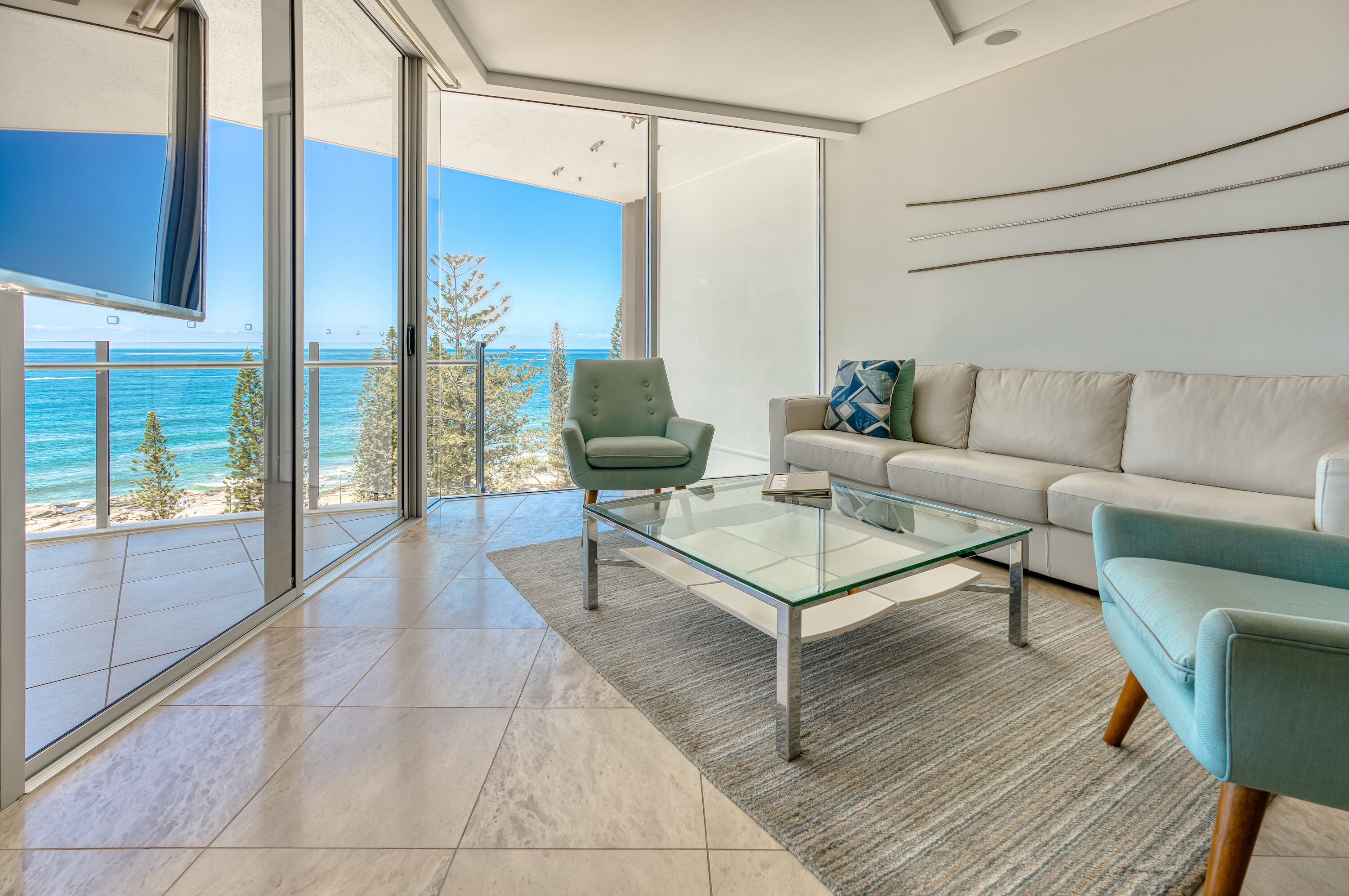 A modern living room with a glass coffee table, a sofa, and a chair, overlooking a beach and ocean.