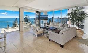A spacious living room with a large sofa, a coffee table, and a dining area with chairs. There is a large window offering a view of the ocean and trees.