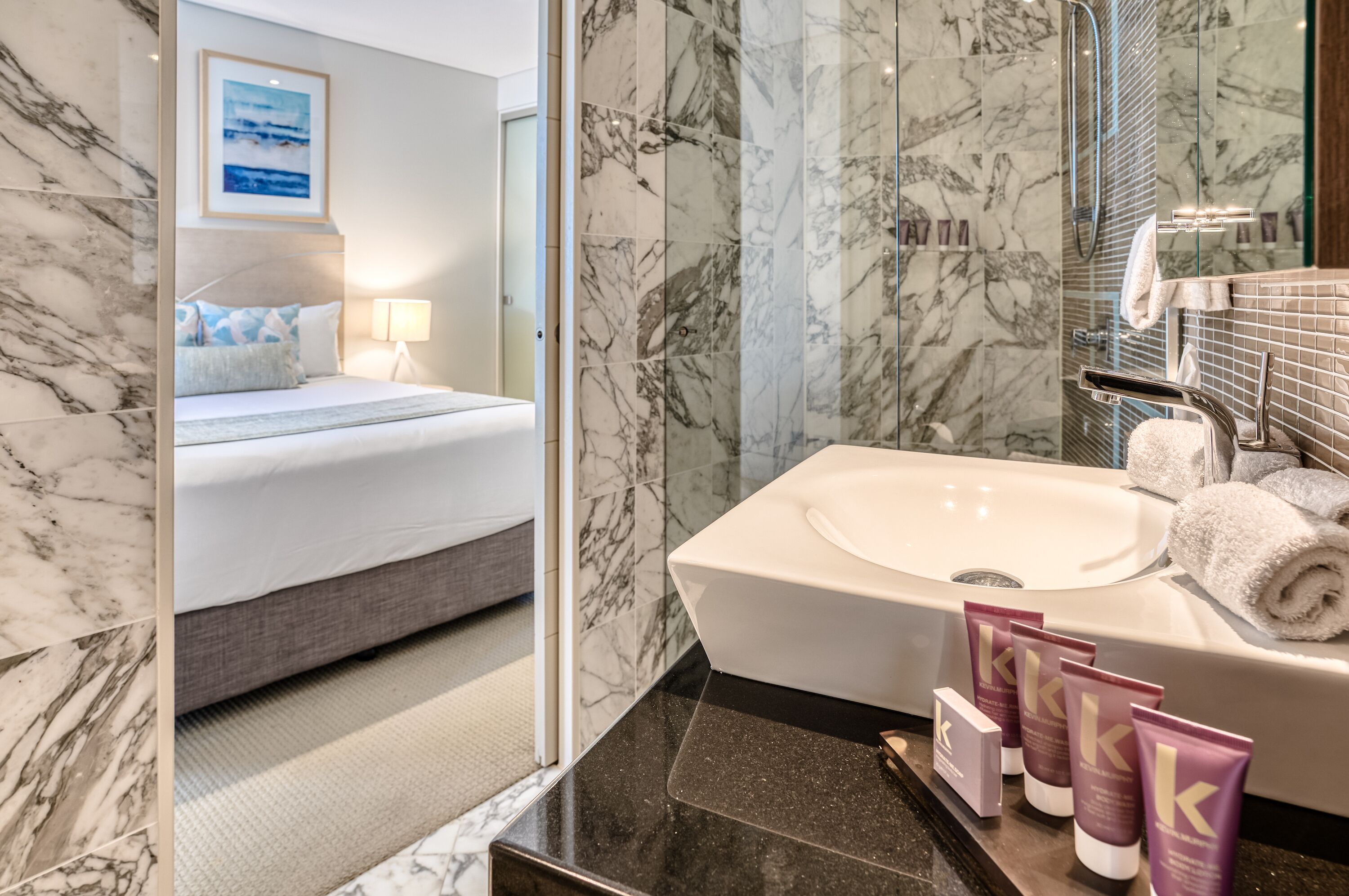 A modern hotel bathroom with a marble-tiled shower, a large sink, and a neatly made bed with a view of the sea through the window.