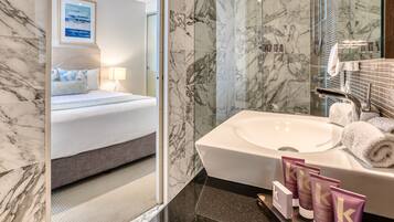 A modern hotel bathroom with a marble-tiled shower, a large sink, and a neatly made bed with a view of the sea through the window.