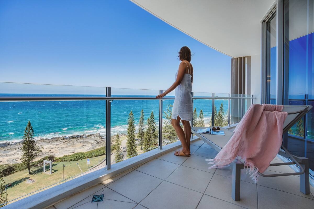 Un balcón con vista despejada al océano y la playa, barandilla de vidrio y una toalla rosa en una silla.