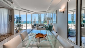 A modern living room with a glass coffee table, beige sofas, and a view of the ocean.