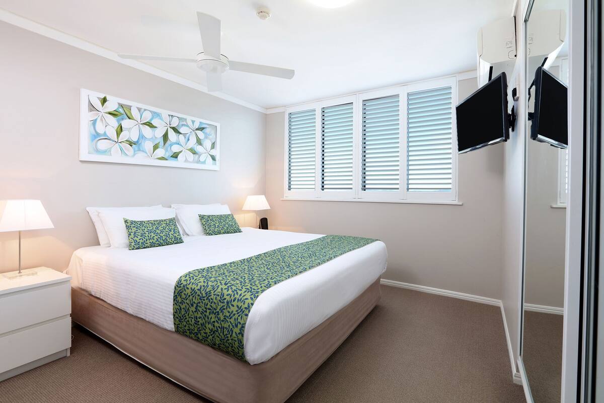 Habitación de hotel con una cama grande, ventilador de techo, televisor de pantalla plana en la pared y ventana con persianas.