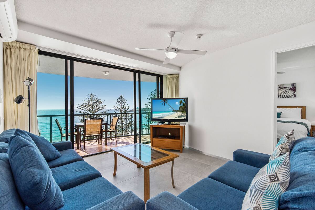 Una sala de estar moderna con un televisor grande, sofás azules, una mesa de centro de vidrio y un balcón con vistas al mar.