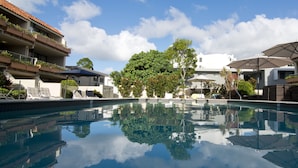 Une piscine entourée de bâtiments dotés de balcons et de verdure, avec des chaises longues et des parasols à proximité.