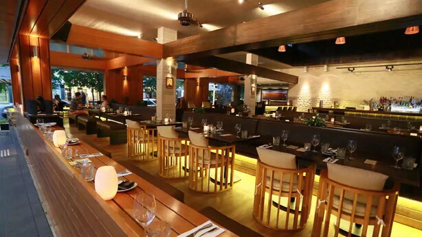 Un restaurant avec de longues tables et des chaises en bois, un coin bar et une vue sur l’extérieur.