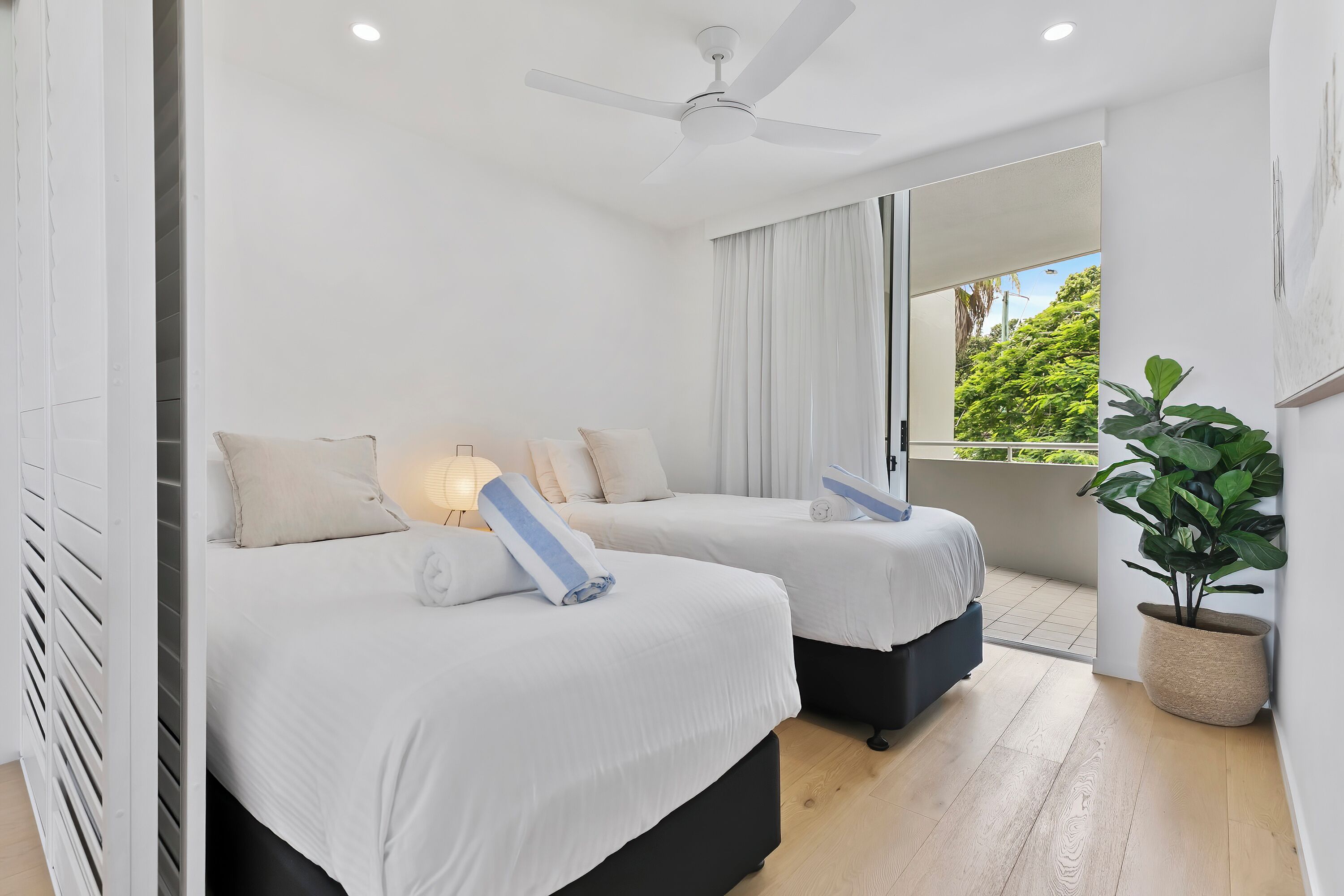 A modern hotel room with two beds, a ceiling fan, a large window with curtains, and a potted plant.