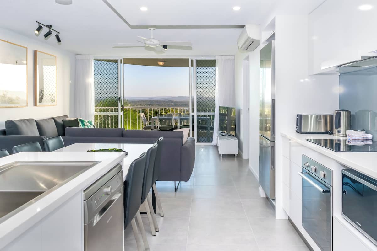 Eine moderne Küche mit eingebautem Ofen, ein Essbereich mit weißem Tisch und Stühlen und ein Balkon mit Blick auf die Landschaft.