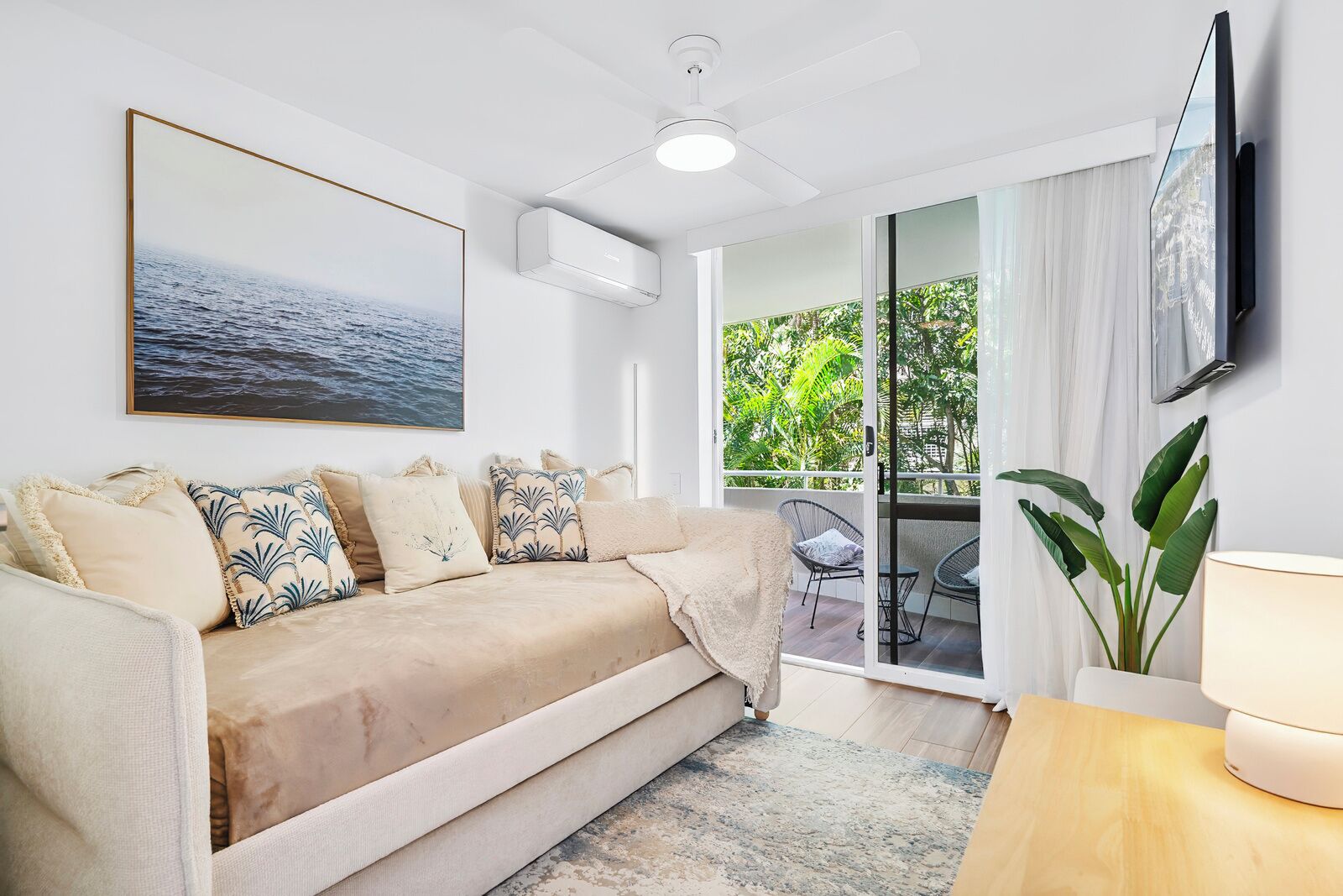 A modern living room with a beige sofa, a flat-screen TV mounted on the wall, and a large window offering a view of greenery.