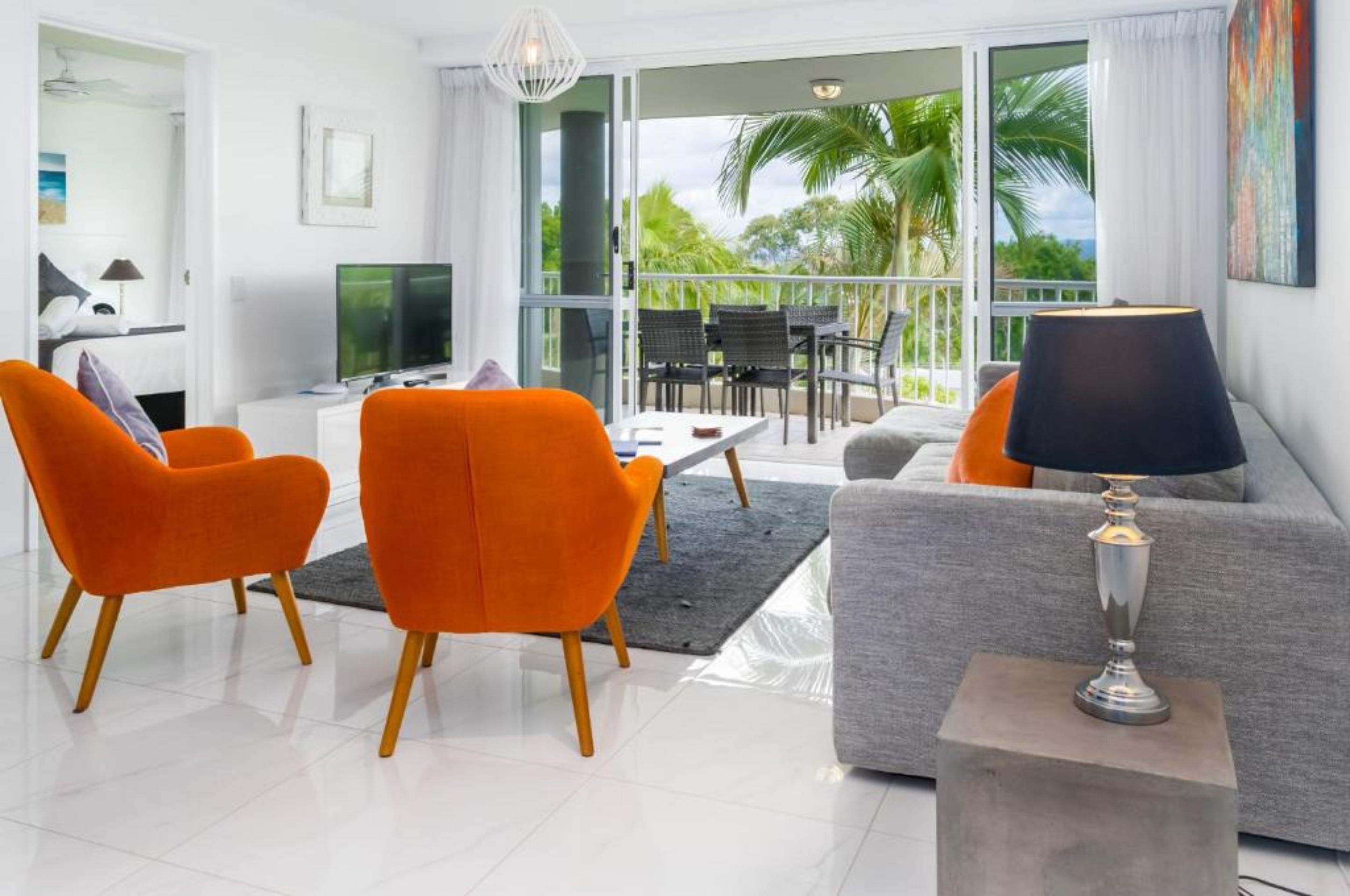 Un salon moderne avec un canapé gris, des fauteuils orange, un téléviseur et un balcon offrant une vue sur des palmiers.