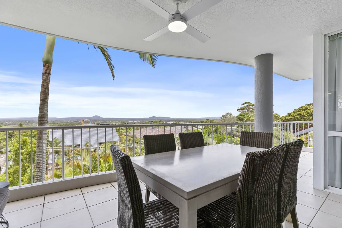 Un balcon avec une table et des chaises, un ventilateur de plafond et une vue sur le paysage urbain et les montagnes.