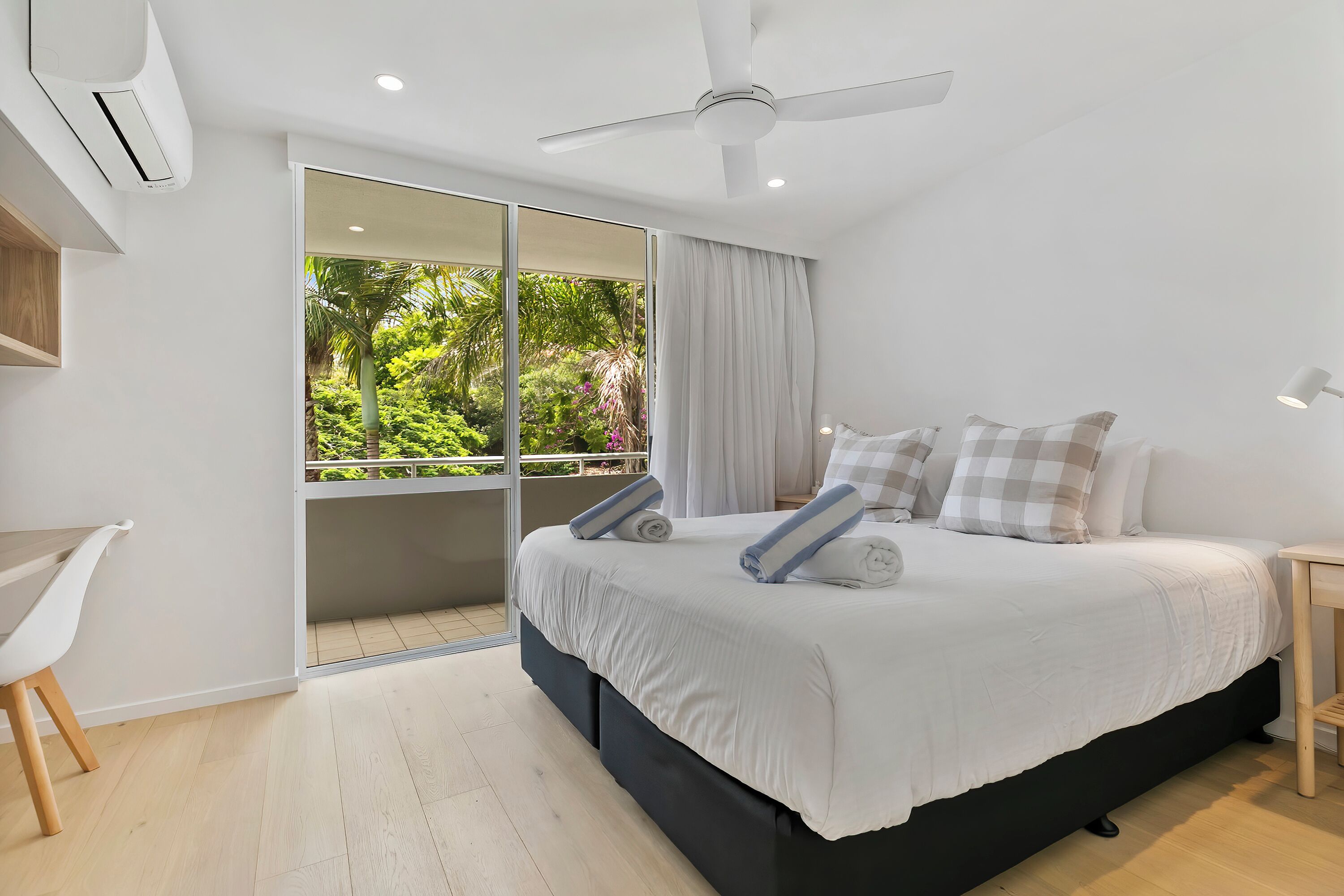 A modern bedroom with a large bed, a desk, and a balcony with a view of greenery.