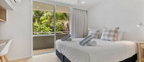 A modern bedroom with a large bed, a desk, and a balcony with a view of greenery.