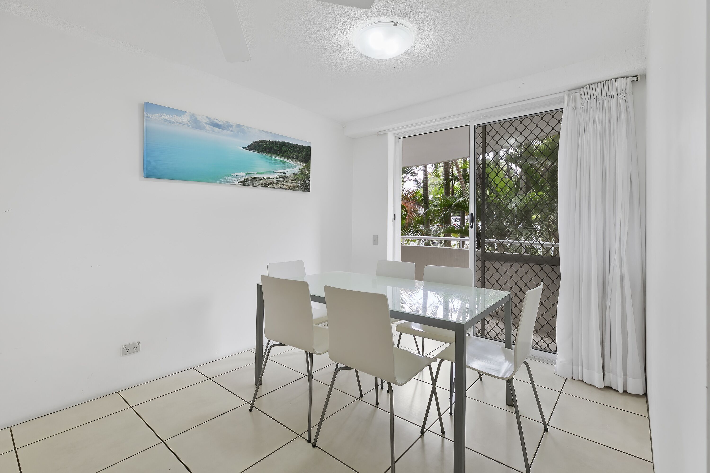 A dining area with a glass table and four chairs, a painting of a coastal landscape, and a view through a sliding glass door to a balcony with a white curtain.