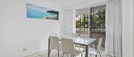 A dining area with a glass table and four chairs, a painting of a coastal landscape, and a view through a sliding glass door to a balcony with a white curtain.