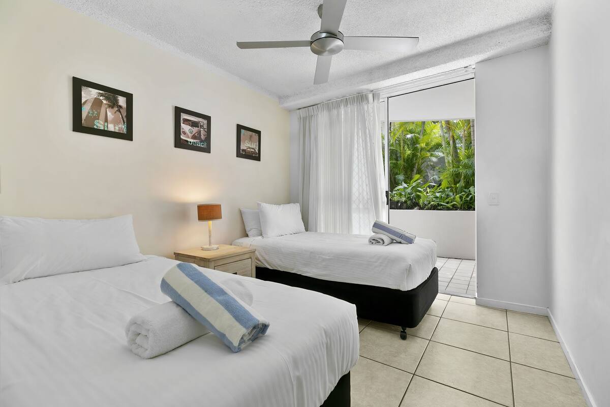 Une chambre d’hôtel avec deux lits, un ventilateur de plafond, une fenêtre avec des rideaux et une vue sur la verdure.