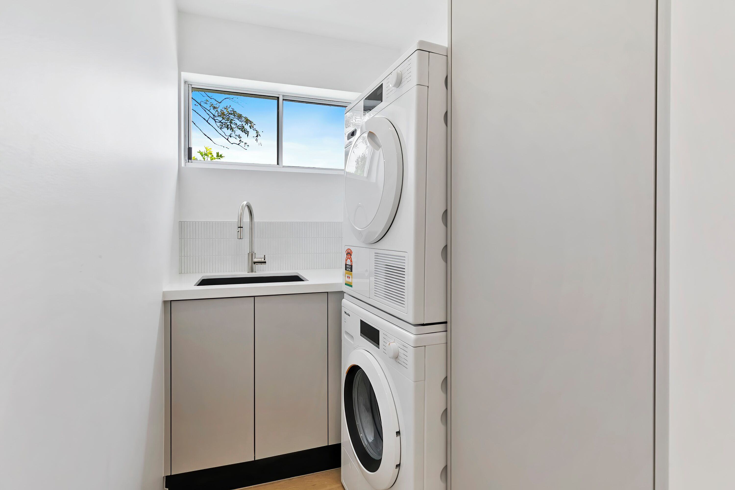 Une petite buanderie équipée d’un lave-linge et d’un sèche-linge superposés, d’une fenêtre offrant une belle vue et d’un évier blanc.