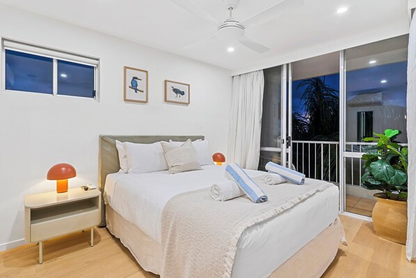 A modern bedroom with a large bed, a nightstand, a ceiling fan, and a view of the outdoors.