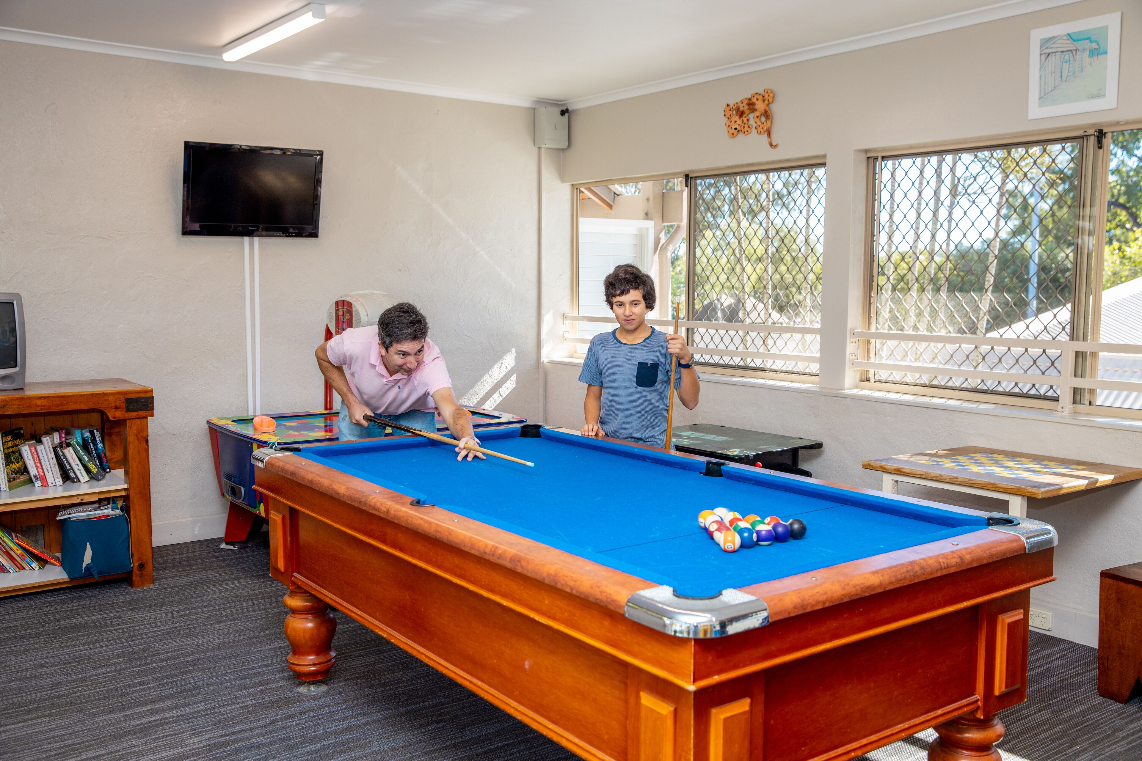 A pool table in a room with a bookshelf, a television, and windows.