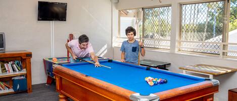 A pool table in a room with a bookshelf, a television, and windows.
