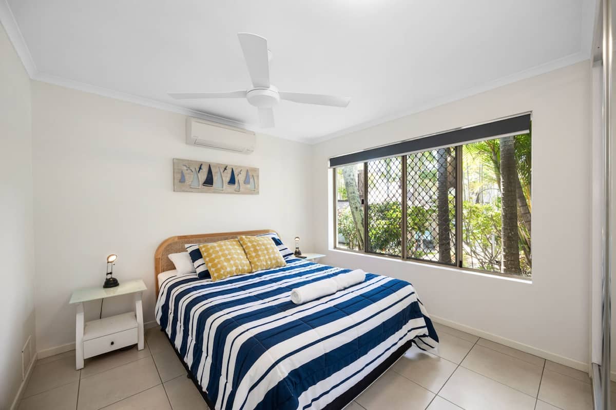 A bedroom with a bed, a nightstand, a ceiling fan, a window with a view of trees, and a wall-mounted air conditioner.